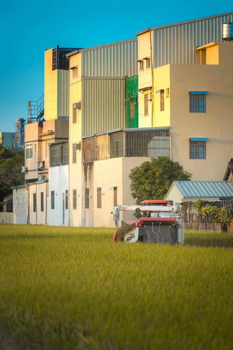 Modern agricultural machinery working in a field near urban residential buildings under clear skies.